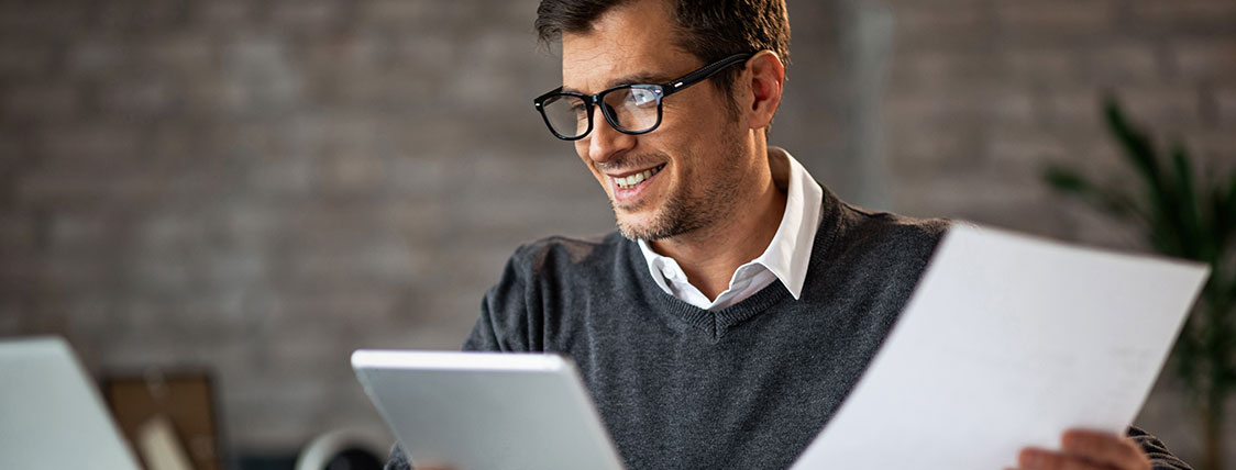 Man at a desk holding a tablet device and piece of paper while looking at a laptop image