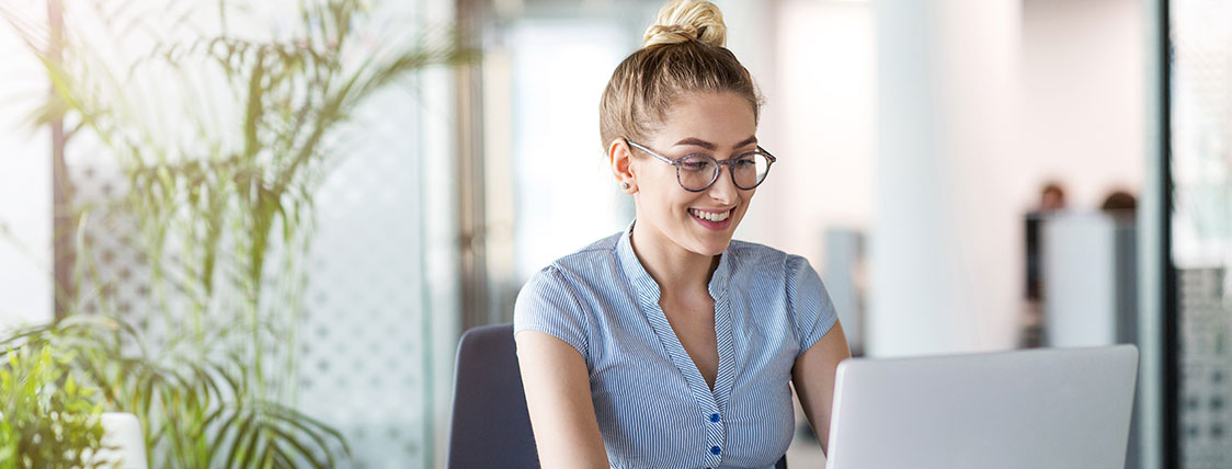 Woman sitting at desk working on a laptop with large plants behind her image