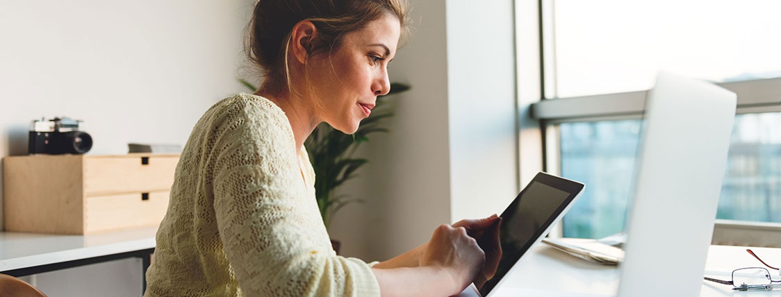 Woman sitting at a desk in front of a computer monitor while looking at a tablet device image