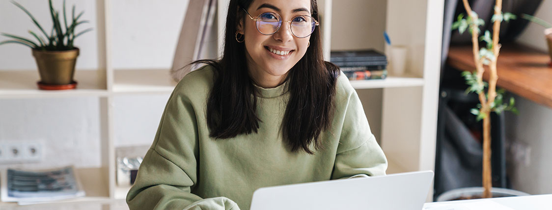 Woman sitting at a desk working on a laptop with a bookcase and plants behind her image