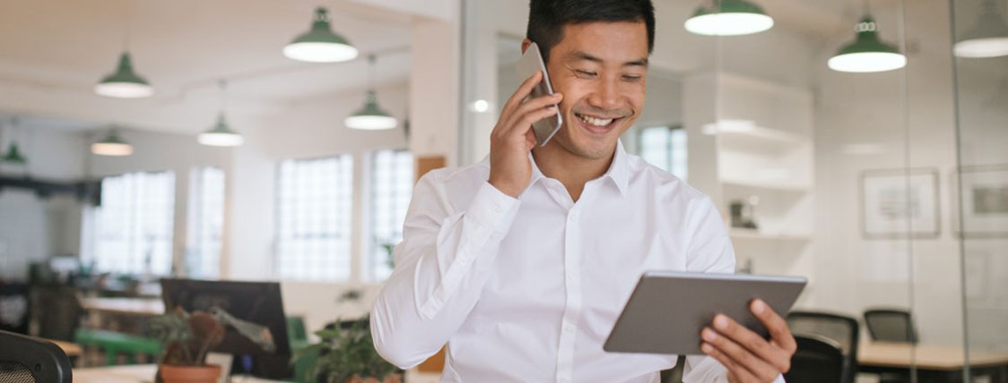 man standing while talking on a cell phone and holding a tablet device in open office area image